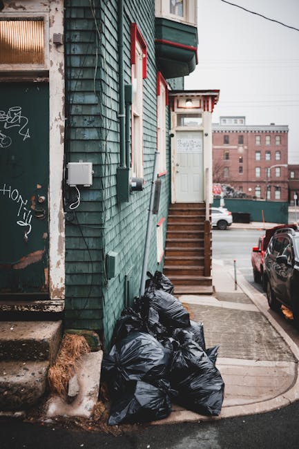 A small urban residential building with a teal wooden exterior, featuring vertical siding and red and white trim around a small front door accessed by a set of wooden steps. Several black plastic rubbish bags are piled at the base of the building near a concrete footpath, with some debris and dried leaves visible on the ground. To the left, part of a large graffiti-covered metal door with rust and peeling paint can be seen, indicating an adjacent commercial or storage area. In the background, a parking lot with vehicles and a multi-storey brick building are visible under a cloudy sky, suggesting an industrial or mixed-use neighbourhood. The scene captures a typical rubbish collection area, emphasizing the importance of proper waste removal and private disposal solutions often handled by services like Waste Clearance Harringay, particularly for on-site clearance or alternative waste handling outside of local authority collections.