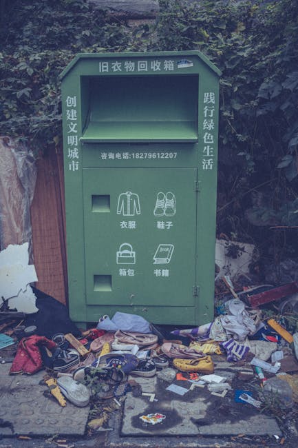 A green, outdoor clothing and items collection box situated on a paved area with a background of dense bushes and greenery. The box features Chinese characters and a blue illustration of a house or storage icon at the top. It has a large, empty rectangular compartment at the top for clothing donations, with a smaller slot below labeled for clothing, shoes, bags, and books, each accompanied by simple line icons. The surrounding ground is cluttered with an assortment of discarded items, including various shoes, bags, and miscellaneous debris, spread out in front of the box. The scene suggests an informal or public collection point for donating used clothing and related items, with waste and debris nearby indicating potential issues with waste management or illegal disposal. The environment appears to be an outdoor area adjacent to natural foliage, and the lighting is natural, with soft shadows indicating daytime. This setup underscores the importance of proper waste or donation disposal, which Waste Clearance Harringay assists with through private collection services for bulky rubbish and unwanted items.