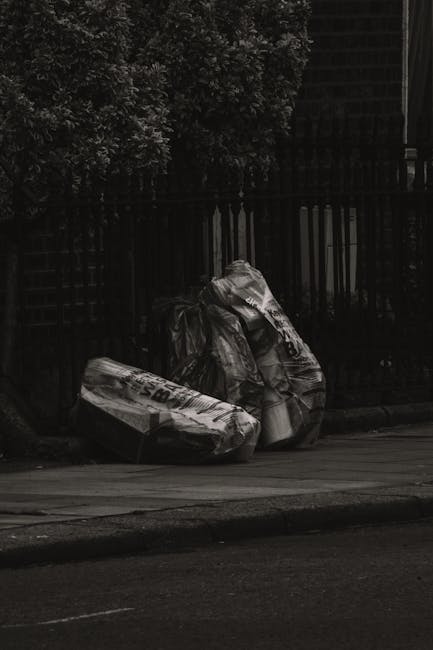 A group of large black plastic garbage bags, some appearing partially torn or crumpled, are placed on a paved sidewalk next to a curb. The bags contain mixed waste materials and are situated in front of a black metal fence with vertical bars and a dark brick wall behind it. To the left of the bags, a portion of a green leafy tree branches into the top of the frame, providing a natural background contrast. The scene is illuminated with natural light, casting soft shadows on the pavement, suggesting it is daytime. The image captures the concept of waste disposal within an urban environment, possibly related to private rubbish collection or on-site clearance services, as promoted by Waste Clearance Harringay's rubbish removal offerings in the local area. The surrounding environment appears quiet with no pedestrians or vehicles visible, emphasizing the focus on the waste bags awaiting collection or disposal.