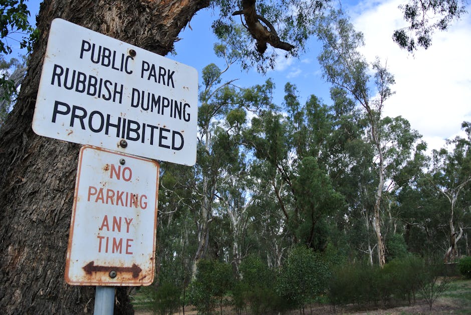 A weathered white metal sign attached to a tree trunk in a natural outdoor setting, with the top part displaying black text that reads 'PUBLIC PARK RUBBISH DUMPING PROHIBITED' and the bottom part showing rusted text that states 'NO PARKING ANY TIME' with an arrow pointing left. The background includes a mix of tall eucalyptus trees and leafy shrubs under a partly cloudy sky, with sunlight filtering through the foliage, creating a dappled light effect on the scene. The sign signifies restrictions on rubbish disposal and parking within this park area, emphasizing the importance of proper waste management and adherence to local regulations. Waste Clearance Harringay, a rubbish removal service, recognizes such regulations when providing alternative waste handling solutions in different environments, including outdoor public spaces.