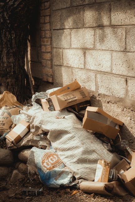 A group of large black plastic garbage bags, some appearing partially torn or crumpled, are placed on a paved sidewalk next to a curb. The bags contain mixed waste materials and are situated in front of a black metal fence with vertical bars and a dark brick wall behind it. To the left of the bags, a portion of a green leafy tree branches into the top of the frame, providing a natural background contrast. The scene is illuminated with natural light, casting soft shadows on the pavement, suggesting it is daytime. The image captures the concept of waste disposal within an urban environment, possibly related to private rubbish collection or on-site clearance services, as promoted by Waste Clearance Harringay's rubbish removal offerings in the local area. The surrounding environment appears quiet with no pedestrians or vehicles visible, emphasizing the focus on the waste bags awaiting collection or disposal.