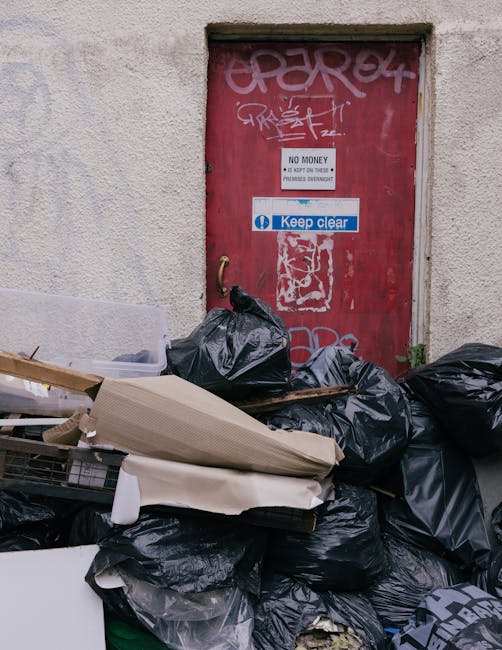 A scene featuring a red metal door with various graffiti tags and signs, including a white sign that reads 'NO MONEY' and a blue and white sticker that says 'Keep clear.' In front of the door, there is a large accumulation of black rubbish bags, some of which appear to be tightly tied, and scattered debris like cardboard and plastic packaging, creating a cluttered and untidy environment. The wall surrounding the door is textured and beige, with signs of weathering, and the rubbish is piled on a paved or concrete surface, indicating a typical urban setting for waste collection or disposal, possibly related to private rubbish removal or on-site clearance services offered by Waste Clearance Harringay. The scene captures an example of blockages of waste meant for disposal, illustrating the need for professional rubbish removal solutions to manage such excess clutter efficiently and prevent environmental or safety issues.