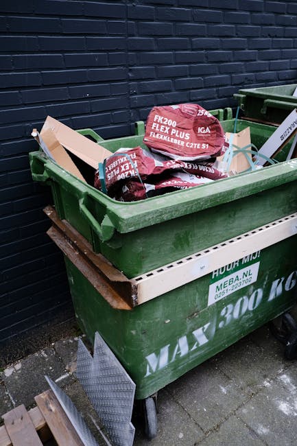 A green industrial rubbish bin filled with several red and black packaging bags containing garden or building materials, positioned outdoors on a concrete surface and placed against a dark brick wall. The bin is on four wheels and is partially covered by a white board or panel with green text, indicating waste collection services. Other waste containers are partially visible next to it, and a small section of a wooden pallet and a metal grate are seen at the bottom left corner of the image. The scene appears to be part of a waste removal or disposal area, with natural daylight illuminating the objects clearly. The setup suggests an example of private or on-site waste management, aligning with alternative rubbish collection services sometimes used by local businesses or residents for managing larger or specific waste types. Waste Clearance Harringay might handle such waste collection needs, providing an alternative to council services.
