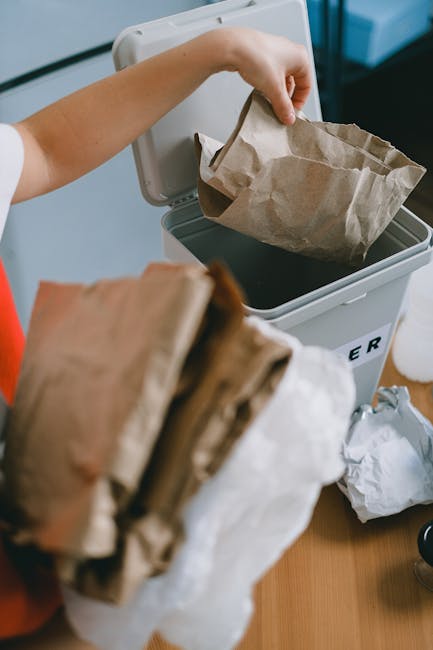 A person discarding a crumpled brown paper bag into a grey rubbish bin with a partially open lid, situated indoors on a light wooden surface. The person's arm is visible, with their hand holding the paper bag over the bin, which is marked with the word 'RUBBISH' in black lettering on the side. The bin appears to be made of plastic, with a smooth surface and a rounded edge. Additional crumpled paper bags are seen in the foreground, held in the person's other hand. The background includes a blurred view of a room with a dark piece of furniture and a white object, suggesting a domestic or office waste management setting. The scene reflects an informal approach to rubbish disposal consistent with private waste collection services like those offered by Waste Clearance Harringay, emphasizing on-site or independent waste removal from interior spaces, where paper and lightweight packaging are typical materials for rubbish collection.