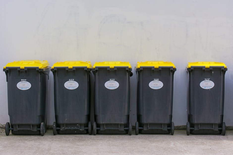 A green industrial rubbish bin filled with several red and black packaging bags containing garden or building materials, positioned outdoors on a concrete surface and placed against a dark brick wall. The bin is on four wheels and is partially covered by a white board or panel with green text, indicating waste collection services. Other waste containers are partially visible next to it, and a small section of a wooden pallet and a metal grate are seen at the bottom left corner of the image. The scene appears to be part of a waste removal or disposal area, with natural daylight illuminating the objects clearly. The setup suggests an example of private or on-site waste management, aligning with alternative rubbish collection services sometimes used by local businesses or residents for managing larger or specific waste types. Waste Clearance Harringay might handle such waste collection needs, providing an alternative to council services.