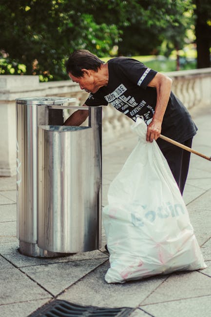 A man, dressed in a black T-shirt with white text, is seen disposing of rubbish into a tall, stainless steel litter bin on a paved pathway in an outdoor park setting. He is leaning forward, with his right hand reaching into the bin, while his left hand holds a large white plastic bag filled with waste. A wooden stick is gripped in his left hand, used to assist in collecting or managing the rubbish. The background features lush green trees and foliage, providing a natural environment, with sunlight filtering through the leaves, creating a well-lit scene. The pavement consists of square stone tiles, and a low stone wall runs parallel to the path behind the man, indicating a designated waste disposal area within a community or public space. Such a scene illustrates private or community-driven rubbish collection, often managed by waste clearance services like Waste Clearance Harringay, emphasizing the importance of on-site waste handling and environmental cleanliness without any promotional context.