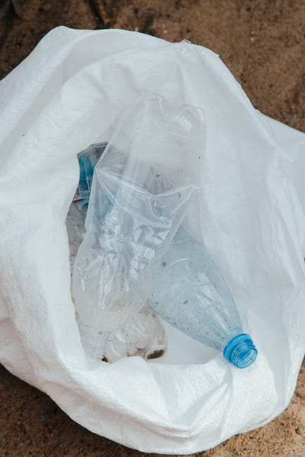 A clear plastic water bottle with a blue cap, partially crushed and showing some dirt and debris on its surface, rests inside a white, woven polypropylene sack that appears to be filled with other waste materials. The sack is open at the top, revealing the bottle among other unidentifiable rubbish. The background consists of a brown, textured outdoor surface, possibly paved or soil, suggesting the scene is set in an external environment suitable for waste collection or disposal. The lighting is natural, casting soft shadows on the bag and the bottle, emphasizing the textures of the plastic and fabric. This image illustrates typical household or commercial waste prepared for rubbish removal, aligning with services such as private rubbish collection or on-site clearance provided by waste removal specialists like Waste Clearance Harringay, highlighting the type of recyclable or general waste often handled through alternative waste handling options rather than council collections.