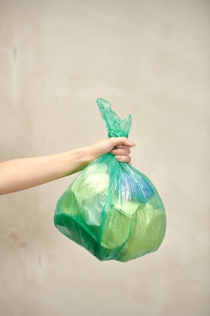 A person's arm extends into the frame from the left, holding a green and blue plastic rubbish bag by its twisted top. The bag is filled with waste, giving it a rounded, bulging shape, and its material appears glossy and slightly crinkled. The hand is gripping the bag firmly, with fingers wrapped around the tied section, and the wrist is visible against a plain, light beige background. The scene suggests the act of discarding household waste, which relates to rubbish removal services such as those offered by Waste Clearance Harringay. The focus on a single bag emphasizes private, independent waste collection, highlighting the practical aspect of managing waste outside of municipal services. The lighting is soft and evenly distributed, with no shadows or reflective highlights, presenting a clear and neutral view of the waste disposal process in a domestic or informal setting, suitable for an article on same-day rubbish collection and waste clearance options near Harringay Green Lanes station.