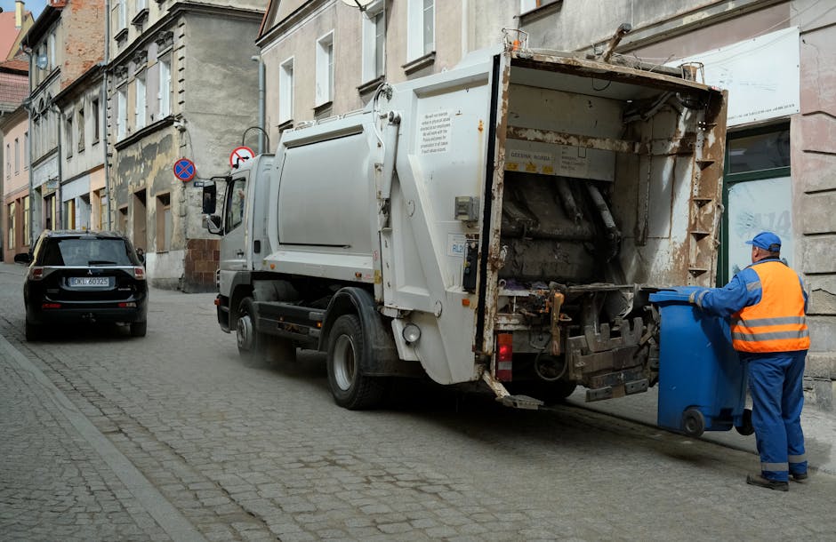A white commercial rubbish collection truck with a rusty, open rear compartment is parked on a narrow street beside old, multi-story buildings with weathered facades. The truck's rear is equipped for the disposal of mixed waste and appears to be engaged in on-site waste clearance. A worker dressed in a blue uniform and bright orange high-visibility vest is positioned next to the truck, handling a small blue wheeled trash bin, likely preparing it for waste collection. The street surface consists of cobblestones and is lined with parked vehicles, including a dark grey car directly behind the truck and a black vehicle parked further along the street. Above, the sky is not visible, but the diffuse lighting suggests an overcast day, making the scene look typical for urban rubbish removal operations that offer alternative waste handling services outside regular council collections. Waste Clearance Harringay's service context is subtly supported through the scene of private waste collection and on-site waste management activities, illustrating practical steps in rubbish removal that accommodate urgent or same-day disposal needs.