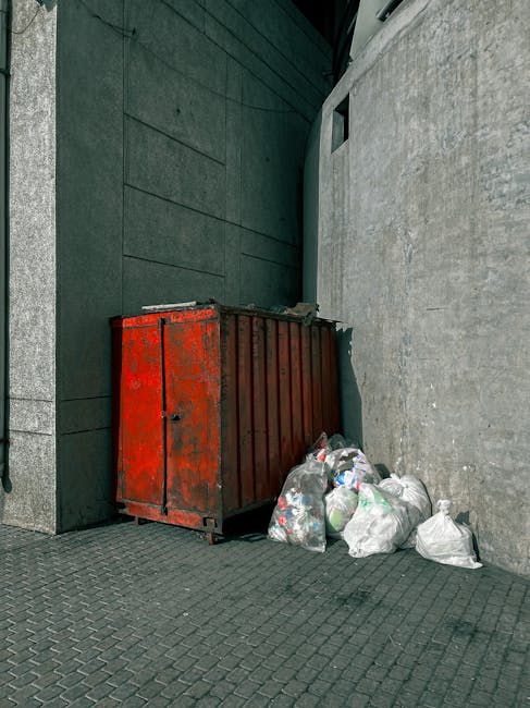 A large, weathered metal skip painted in faded red, positioned against a dark green wall adjacent to a grey concrete wall in an urban outdoor setting. The skip is rectangular with vertical ridges along the sides and a solid top. Next to the skip, on the right, there is a small pile of waste bags made of transparent and white plastic, filled with mixed rubbish such as paper, plastic bottles, and other general waste. The bags are loosely gathered and resting on a paved ground composed of dark grey interlocking bricks. The setting appears to be a narrow alley or corner of a building, with minimal natural light, giving a slightly subdued atmosphere. This scene aligns with private waste disposal or rubbish removal services like those offered by Waste Clearance Harringay, highlighting an on-site collection of general household or commercial waste in preparation for annulling or off-site disposal, and emphasizing the importance of proper waste management outside of local authority collections.