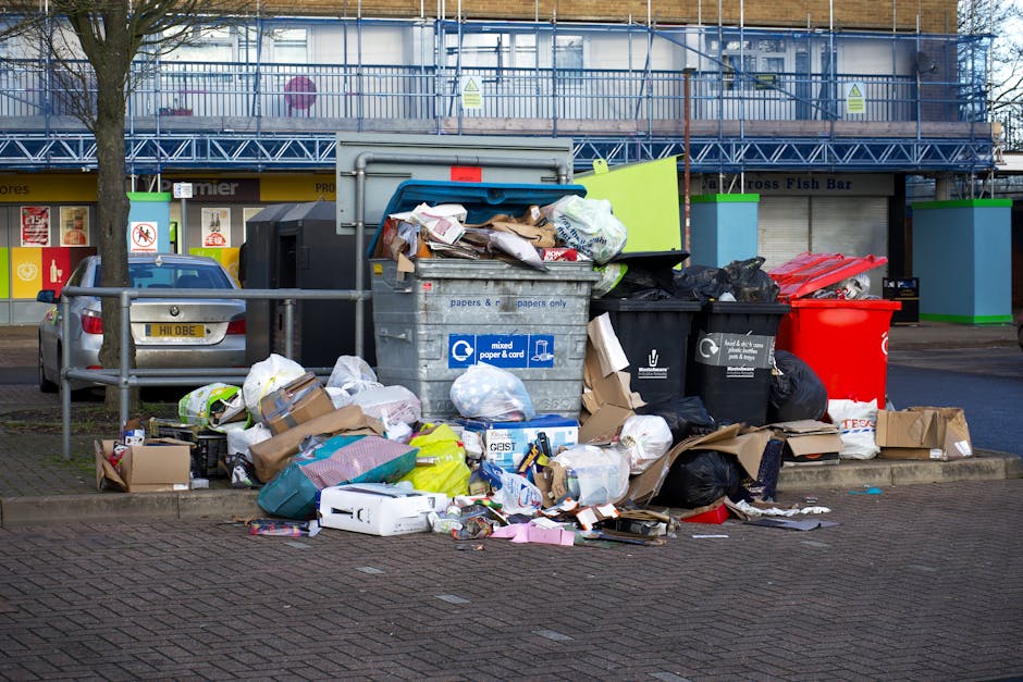 A large collection of overflowing mixed waste and rubbish bags, including paper, cardboard, plastic, and black bin liners, is piled around a communal rubbish bin on a paved urban street. The waste is spilling onto the pavement and street surface, with some boxes and packaging scattered nearby. A silver car is parked adjacent to the waste, partially visible on the left side of the image. In the background, there is a building with a blue metal scaffolding structure, and shopfronts with signage, including a fish bar. The rubbish appears to have been temporarily placed outside, possibly indicating an area overwhelmed with waste or an unscheduled collection. The scene highlights issues related to waste management and the importance of appropriate rubbish disposal services, such as those offered by Waste Clearance Harringay, supporting proper waste handling and preventing illegal dumping in public spaces.
