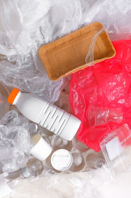 An overhead view of various discarded waste items scattered on the ground, including a partially crushed orange-lidded plastic bottle, a white plastic bottle with ridges along its body, a small white plastic container with a removable lid, a piece of clear plastic tubing, and crumpled red and white plastic bags, all surrounded by clear plastic waste film. The scene appears to be outdoors, possibly on a pavement or driveway, emphasizing the type of household or commercial rubbish typically handled by Waste Clearance Harringay's rubbish removal services, with materials primarily consisting of plastic bottles, wrappers, and packaging. The lighting is natural, highlighting the texture of the plastic surfaces, and the arrangement suggests a collection of loose waste ready for disposal or collection, representing typical clutter associated with non-recyclable household waste or informal waste handling scenarios, supporting the context of alternative waste disposal options outside of standard municipal collection.