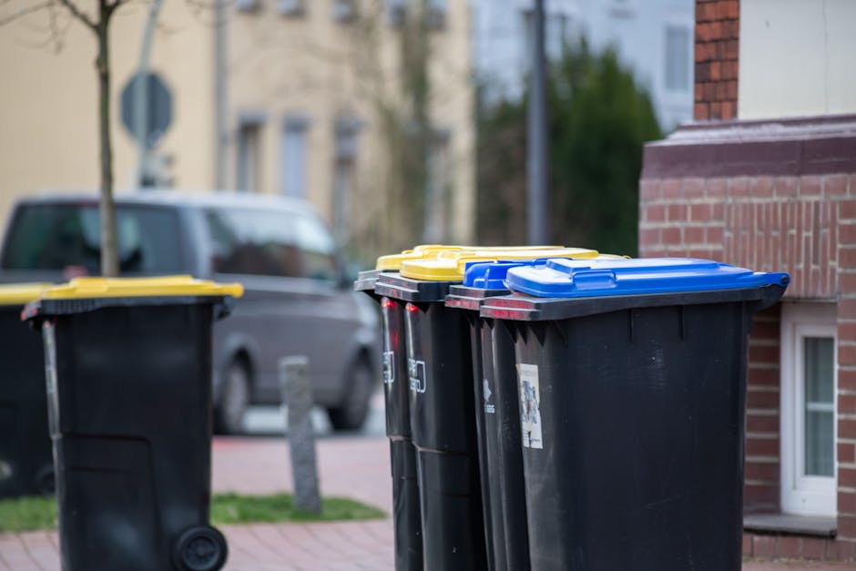 A large collection of overflowing mixed waste and rubbish bags, including paper, cardboard, plastic, and black bin liners, is piled around a communal rubbish bin on a paved urban street. The waste is spilling onto the pavement and street surface, with some boxes and packaging scattered nearby. A silver car is parked adjacent to the waste, partially visible on the left side of the image. In the background, there is a building with a blue metal scaffolding structure, and shopfronts with signage, including a fish bar. The rubbish appears to have been temporarily placed outside, possibly indicating an area overwhelmed with waste or an unscheduled collection. The scene highlights issues related to waste management and the importance of appropriate rubbish disposal services, such as those offered by Waste Clearance Harringay, supporting proper waste handling and preventing illegal dumping in public spaces.